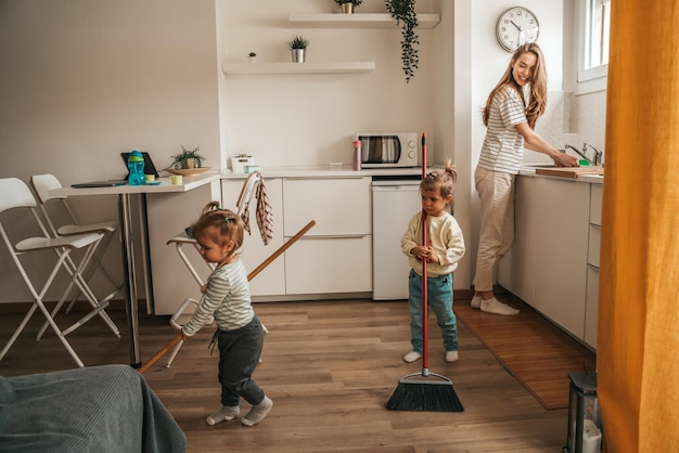 笑顔の幸せな若い母親が皿を洗い、子供たちがモップで床を掃除しているのを見て