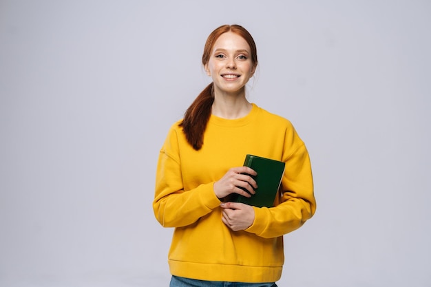 Smiling happy young woman college student yellow sweater\
holding book on isolated gray background