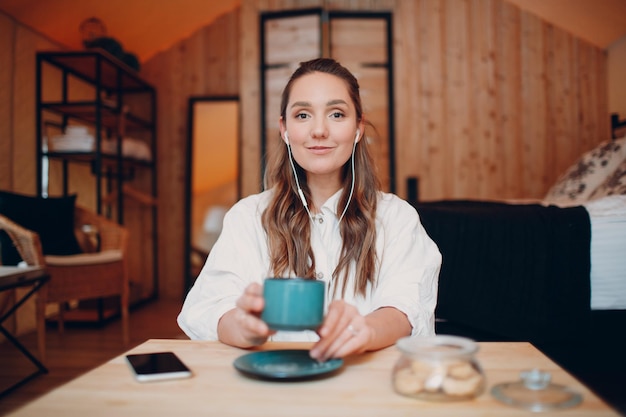 Smiling happy young woman sitting at table at home behind\
computer laptop and talking on video call girl female with cup of\
tea or coffee speaking online on webcam indoors