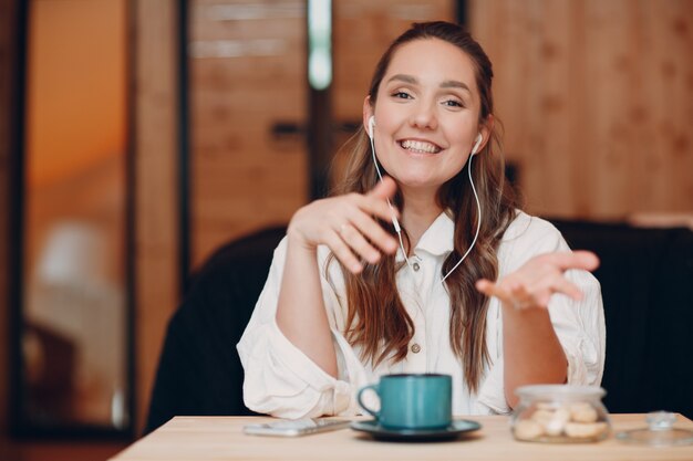 Smiling happy young woman sitting at table at home behind computer laptop and talking on video call ...