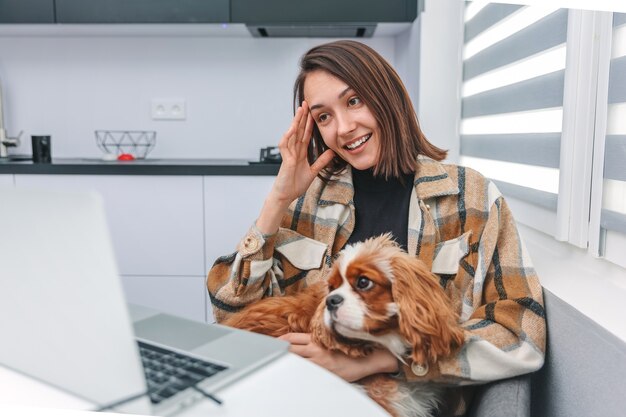 집에 앉아 노트북을 보고 있는 강아지와 함께 웃고 있는 행복한 젊은 여성