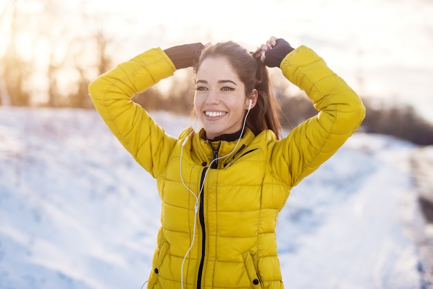 smiling hardworking fitness girl with earphones in winter sportswear tying a ponytail outside in snow nature.