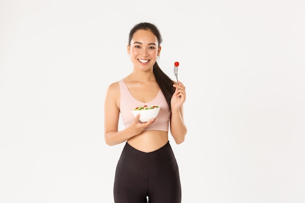 Улыбающаяся здоровая и стройная азиатская девушка в спортивной одежде, держащая салат и собирающая помидор на вилке, оставаясь в форме с помощью диеты и специальной тренировки.