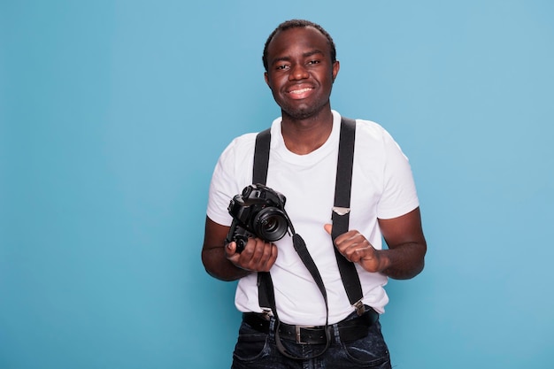 Sorridente fotografo professionista cordiale con fotocamera dslr mentre si trova su sfondo blu. giovane adulto sicuro e bello che indossa abiti alla moda pur avendo un dispositivo fotografico.