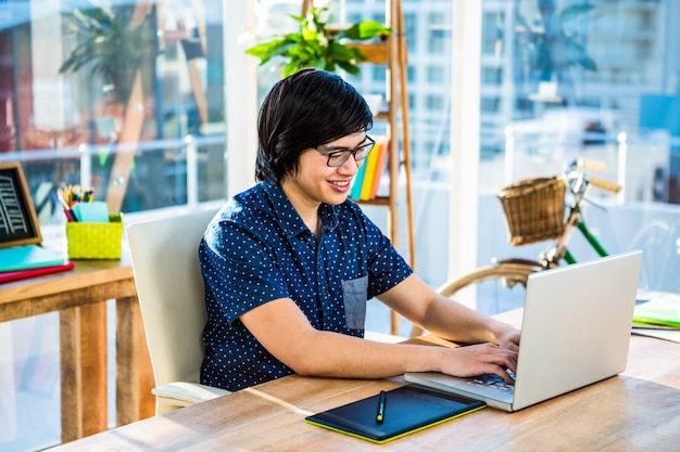 Smiling hipster businessman using laptop and graphic tablet in office