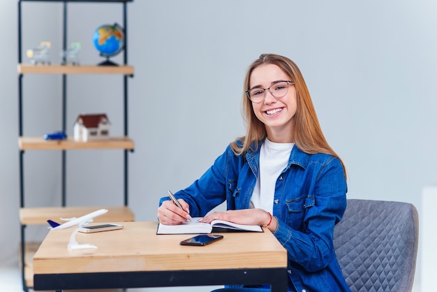 Smiling hipster girl in eyeglasses works and makes some notes and looking at camera.