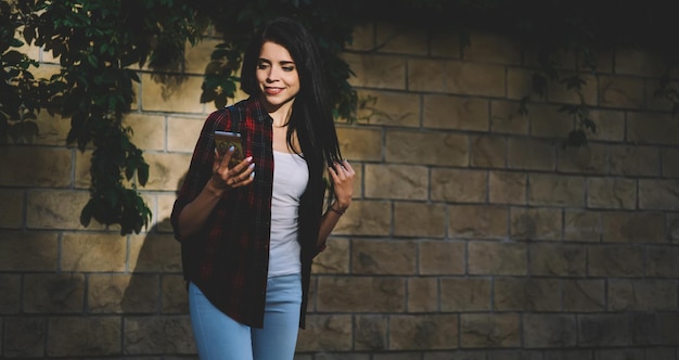 Smiling hipster girl holding modern telephone in hand while checking mail outdoors at summer day