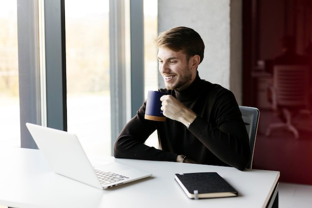 Smiling hipster guy holding cup in his hand sitting at desk with laptop Man working in the office