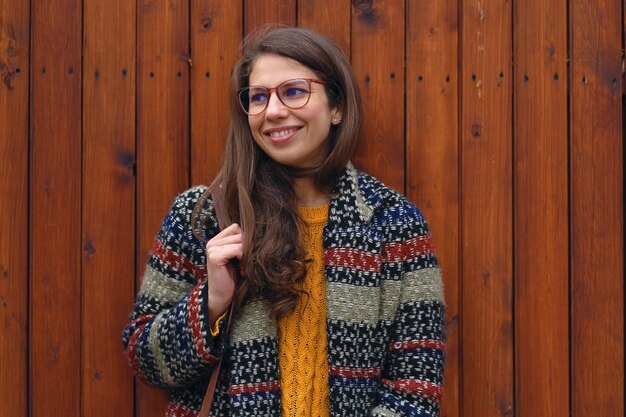 Smiling hipster student girl wearing colorful coat and sweater posing on wood wall. Looking to copy space on the right.