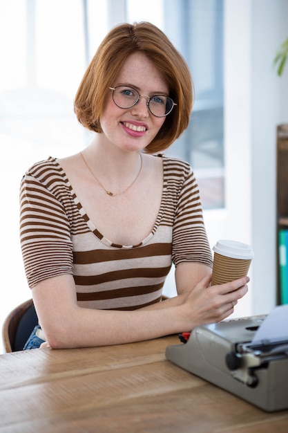 smiling hipster woman holding a coffee cup, in front of her typewriter