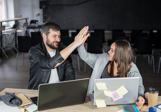 Smiling hipster young couple using laptops and giving high five in the office or lecture hall.