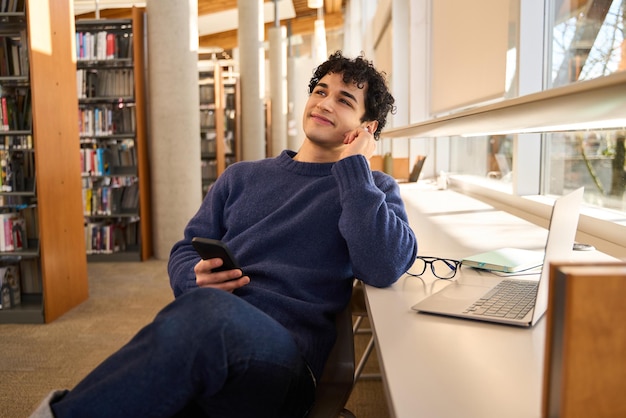 Улыбающийся латиноамериканец с мобильным телефоном сидит за столом с ноутбуком в кампусе библиотеки
