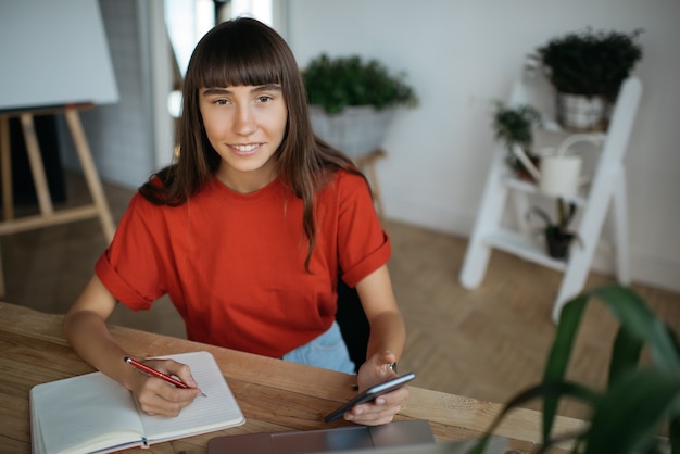 Smiling hispanic woman using mobile phone, taking notes, working from home