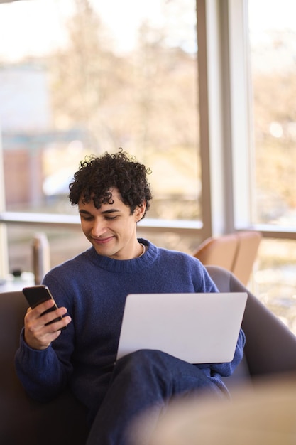 Улыбающийся латиноамериканский молодой человек с помощью мобильного телефона, работая удаленно на ноутбуке в офисе