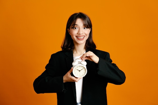 Smiling holding alarm clock young beautiful female wearing black jacket isolated on orange background