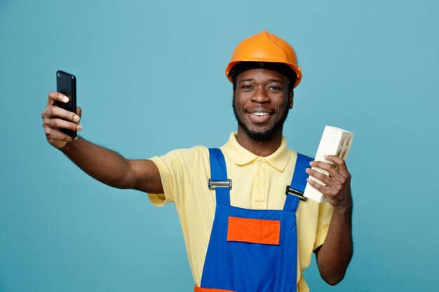 Smiling holding brick young african american builder in uniform take a selfie isolated on blue background