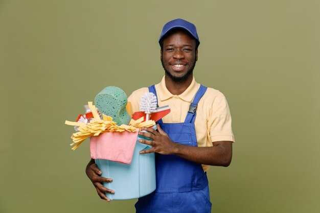 Smiling holding bucket of cleaning tools young africanamerican cleaner male in uniform with gloves isolated on green background