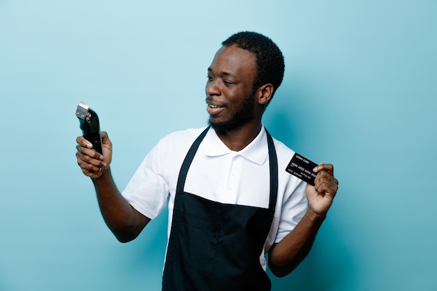Улыбающийся держащий карточку с машинкой для стрижки волос молодой африканский парикмахер в униформе, изолированный на синем фоне