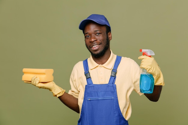 Улыбаясь, держа чистящее средство губкой, молодой афроамериканец-уборщик мужчина в униформе с перчатками, изолированными на зеленом фоне