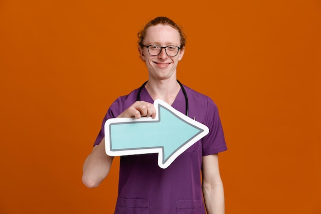 Smiling holding direction mark young male doctor wearing uniform with stethoscope isolated on orange background