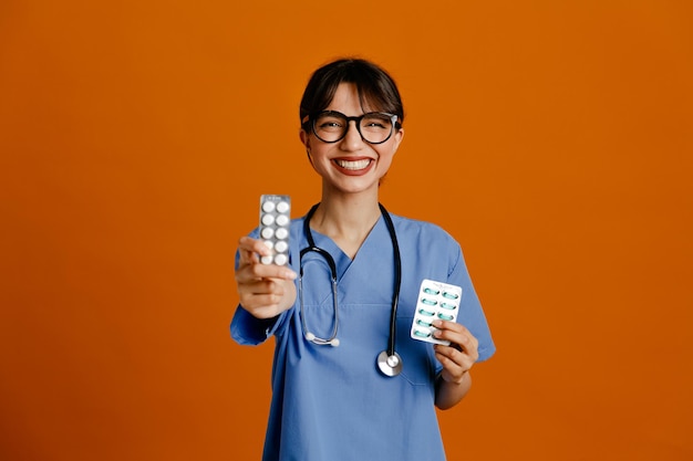 Smiling holding pills young female doctor wearing uniform fith stethoscope isolated on orange background