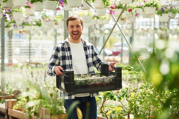 Foto sorrido che tiene una scatola di plastica con piante florista che lavora in un centro giardino un impiegato di successo è in una serra luminosa