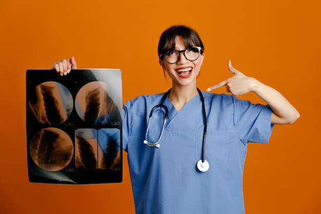 Smiling holding and points at xray young female doctor wearing uniform fith stethoscope isolated on orange background