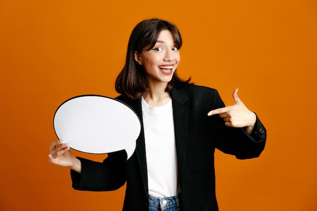 Smiling holding speech bubble young beautiful female wearing black jacket isolated on orange background