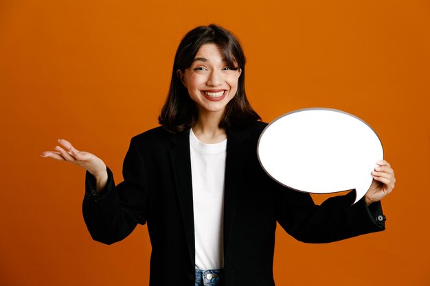 Smiling holding speech bubble young beautiful female wearing black jacket isolated on orange background