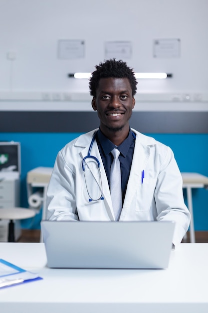 Smiling hospital doctor specialist sitting at clinic cabinet desk using work laptop. Polyclinic medic sitting at office desk in healthcare facility workspace while using computer to check patient repo