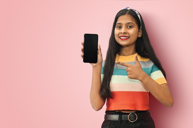 Smiling Indian Asian girl posing isolated is holding blank screen mobile phone and pointing finger to a copy space