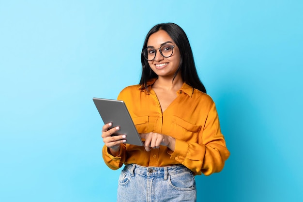 Smiling indian lady using digital tablet wearing eyeglasses blue background