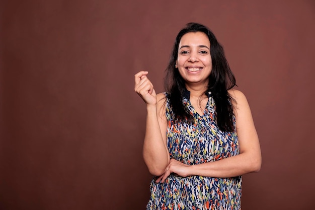 Smiling indian woman posing portrait, happy lady holding hand folded, snapping fingers. Cheerful person standing, looking at camera, front view studio medium shot on brown background