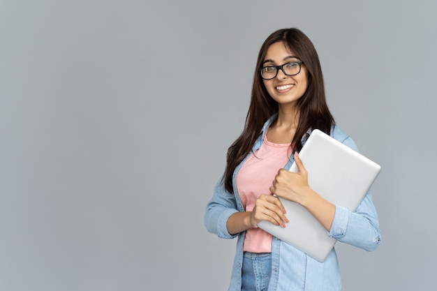 灰色の背景に隔離されたラップトップを保持しているインドの若い女性学生の笑顔