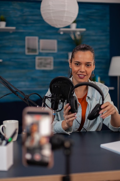 Smiling influencer recording podcast with headphones and microphone. Vlog headset review in front of recording smartphone. Content creator in home studio filming video for social media.