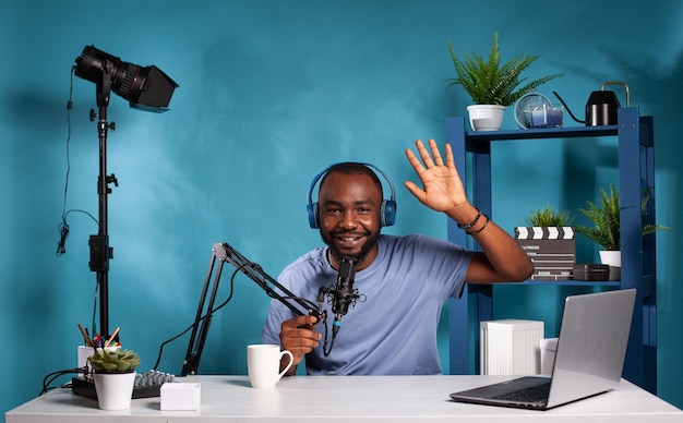 Smiling influencer wearing wireless headphones waving hello in recording studio with professional microphone and video light. Happy vlogger doing hand gesture in front of audio podcast setup.