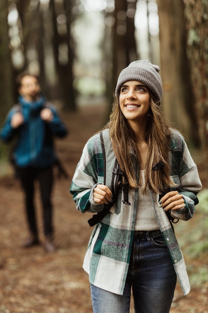 Smiling inspired young european guy and lady tourists in jackets hat with backpack walk in autumn forest