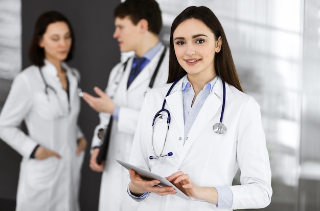 Smiling intelligent woman-doctor is holding a tablet computer in her hands, while she is standing together with her colleagues in a clinic. Physicians at work. Perfect medical service in a hospital. M