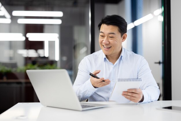 Photo smiling japanese male entepreneur having online video call on modern laptop while holding notebook and pen in hands positive man making notes during conversation with interlocutors at office