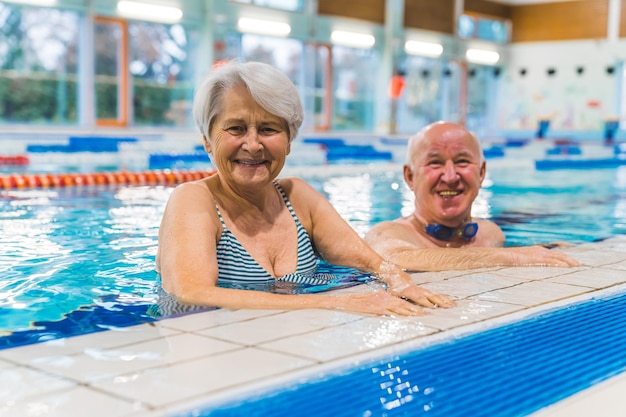 Smiling and joyful aged couple at the side of the indoor pool looking at the camera