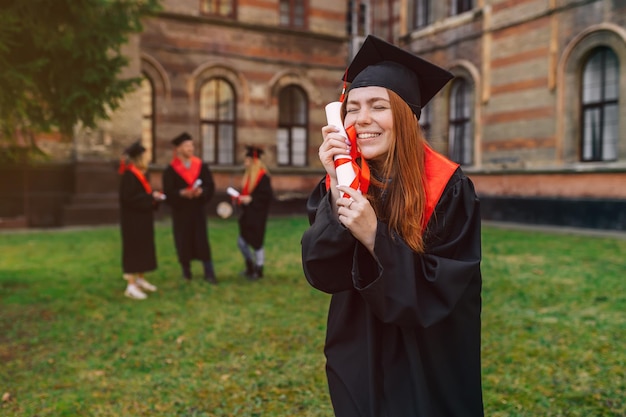 Smiling joyful redhaired girl hugging a diploma in her hands\
student in graduation gown and peaked cap