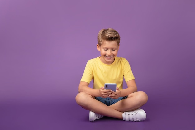 웃고 있는 어린 소년이 휴대폰을 보고 게임을 하는 비디오를 보고 있습니다. 쾌활한 아이는 보라색 배경 스튜디오 샷에 바닥에 앉아 휴대폰을 사용합니다