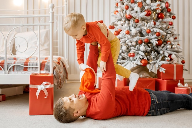 Smiling kid boy in red sweater flying in his fathers arms with happy funny face on the background of the New Year Tree and Christmas gifts