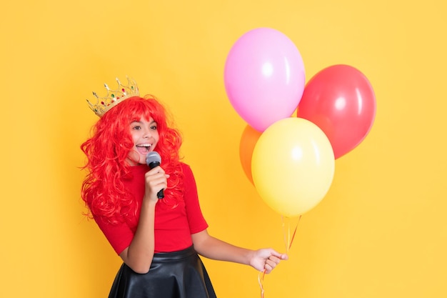 Smiling kid in crown with microphone and party balloon on yellow background