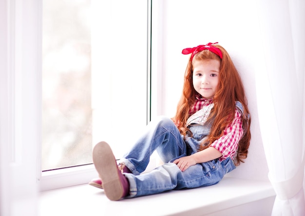 Smiling kid girl 3-4 year old sitting on windowsill in room Looking at camera Childhood