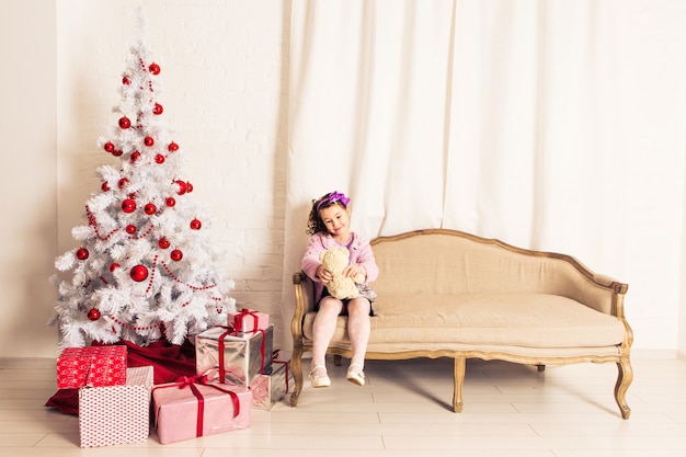 Smiling kid girl holding white teddy bear with christmas tree indoors. Childhood.