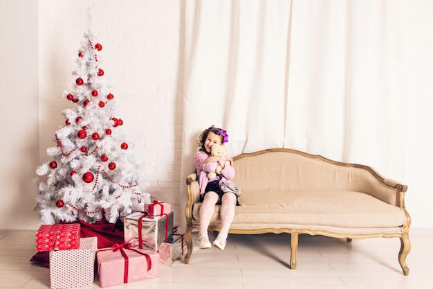 室内のクリスマスツリーと白いテディベアを保持している子供の女の子の笑顔。子供時代。