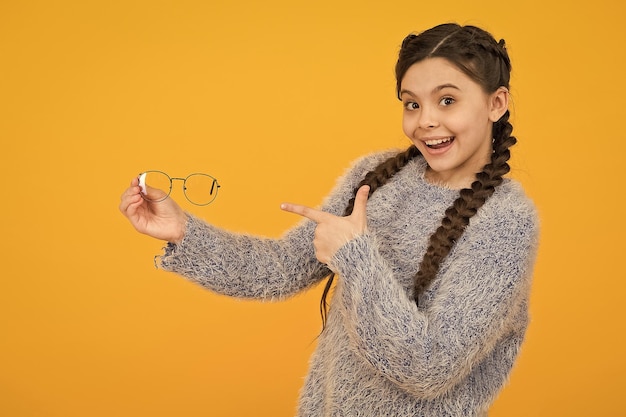 Smiling kid girl with stylish braided hair pointing finger on eyeglasses on yellow background healthcare