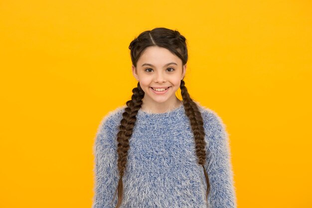 黄色の背景のニットにスタイリッシュな編んだ髪を持つ笑顔の子供女の子