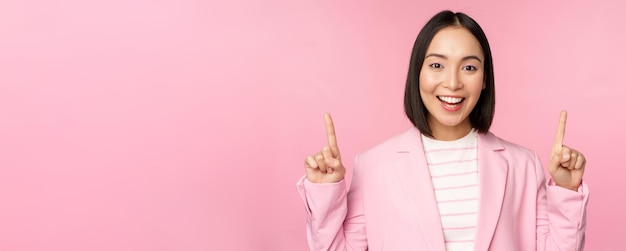 Smiling korean businesswoman pointing fingers up showing advertisement banner or logo on top standing in suit over pink background